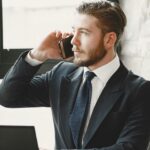 Professional businessman in suit making phone call at desk with laptop in office environment.